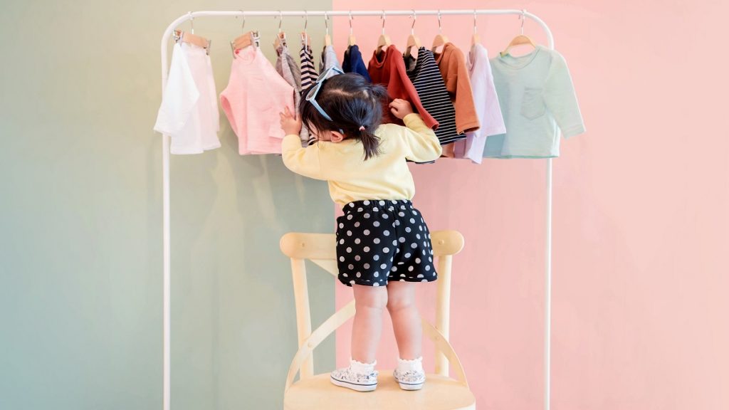 Soft Focus Of A Two Years Old Child Choosing Her Own Dresses From Kids Cloth Rack